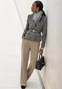 Graham zweireihiger Blazer mit silbernen Knöpfen, beige weit geschnittene Hose, grauer Schal und eine schwarze Handtasche mit goldenen Nieten.