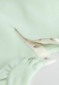 Vêtement vert clair avec une texture douce, fermetures à pression et bords volantés ; présente un tissu intérieur contrastant avec un motif floral.