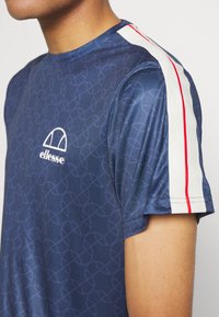 Granatowa koszulka sportowa z geometrycznym wzorem, z białym logo i czerwono-białymi paskami na rękawach. Miękki materiał, krótkie rękawy.