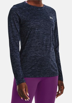 Frau trägt ein marineblaues, langärmeliges Sportshirt mit kleinem Logo auf der Brust und lila Leggings, stehend vor weißem Hintergrund.