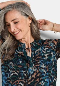 Blusa multicolore in tessuto leggero con motivo floreale nei toni del verde, blu e marrone; scollatura elasticizzata con dettaglio a fiocco.