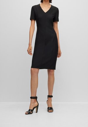 Mujer que lleva un vestido negro ajustado hasta la rodilla con mangas cortas y sandalias negras de tacón alto con correas en el tobillo.