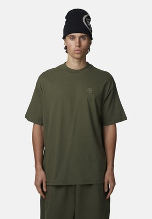 Giovane uomo che indossa una t-shirt oversize verde oliva e pantaloni abbinati, con un berretto nero con un logo bianco.