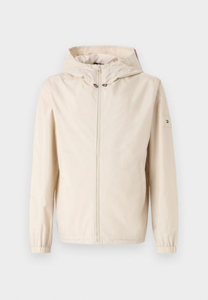 Beige lett jakke med hette, elastiske mansjetter og glidelås foran. Har et logo-patch på venstre erme og en glat tekstur.