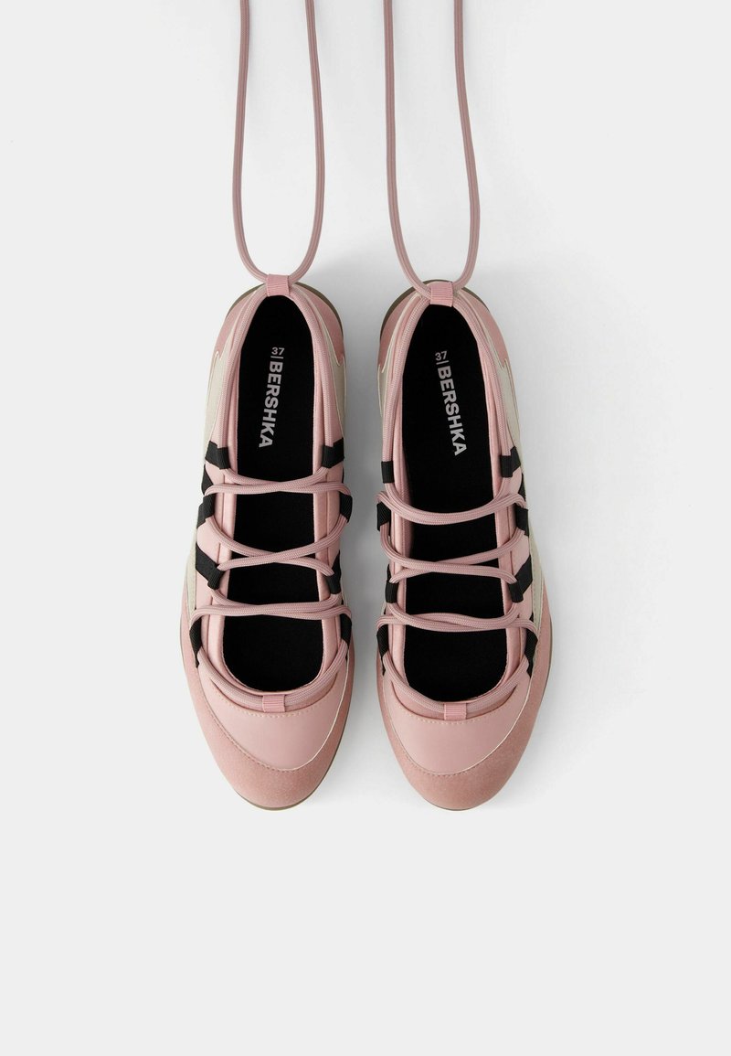 Un par de zapatillas Bershka de color rosa claro con acentos en negro y beige, que presentan cordones en cruz y cordones en bucle colgando arriba, sobre un fondo blanco.