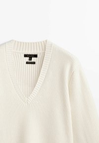 Pull en tricot couleur crème avec un décolleté en V et une texture côtelée. Étiquette indiquant la taille médium, fabriqué en Turquie. Design classique avec des détails subtils.