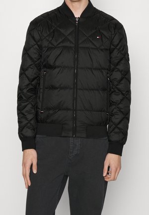 Hombre con chaqueta bomber negra acolchada con cremallera frontal y bolsillos con cremallera, combinada con pantalones oscuros.