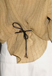 Tessuto oro chiaro con texture, dotato di un laccio nero annodato, con aglette dorate, che si drappeggia su un tessuto bianco. Focus sui dettagli del bordo inferiore e della raccolta.