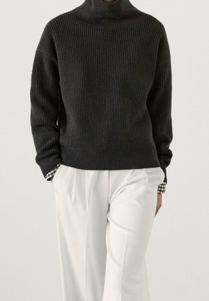 Personne portant un pull à col roulé côtelé foncé sur une chemise à carreaux noir et blanc, associé à un pantalon blanc taille haute.