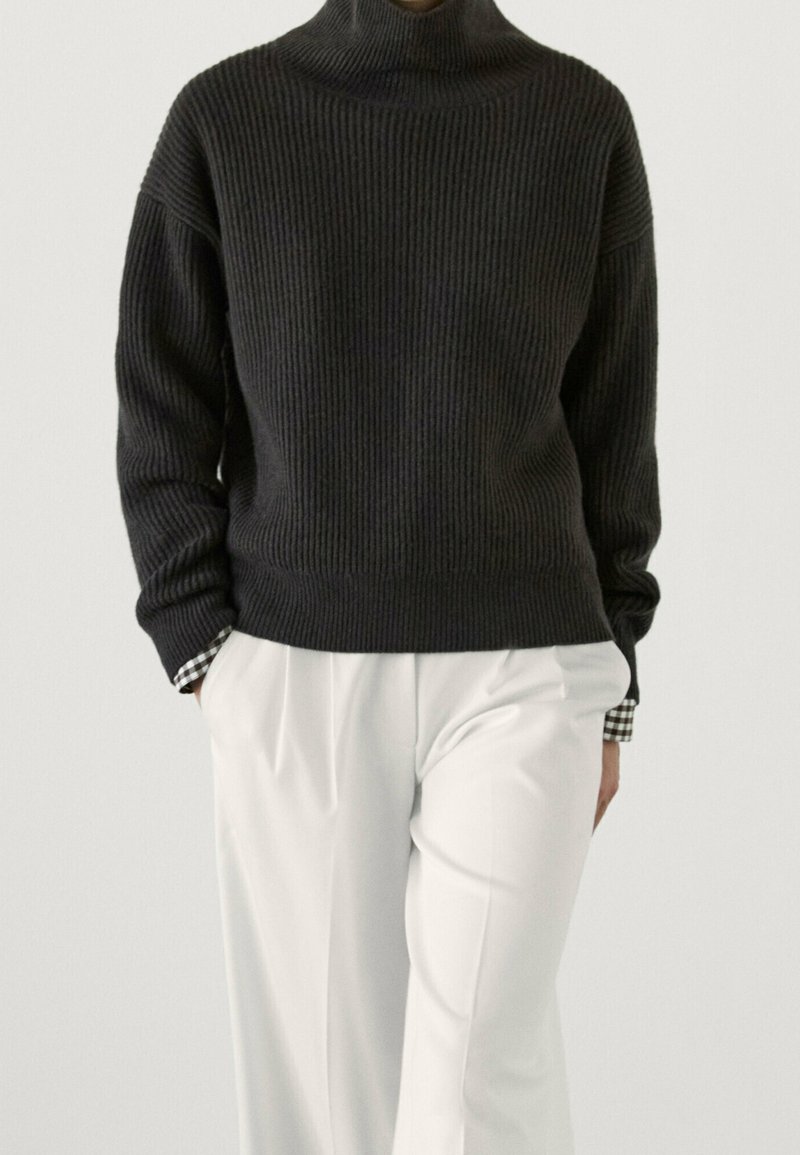 Personne portant un pull à col roulé côtelé foncé sur une chemise à carreaux noir et blanc, associé à un pantalon blanc taille haute.