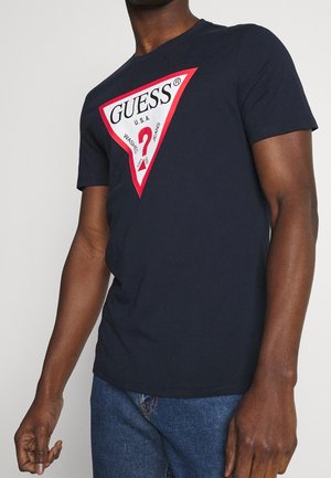 Mann trägt ein marineblaues T-Shirt mit einem großen roten und weißen dreieckigen GUESS-Logo und blauen Jeans.