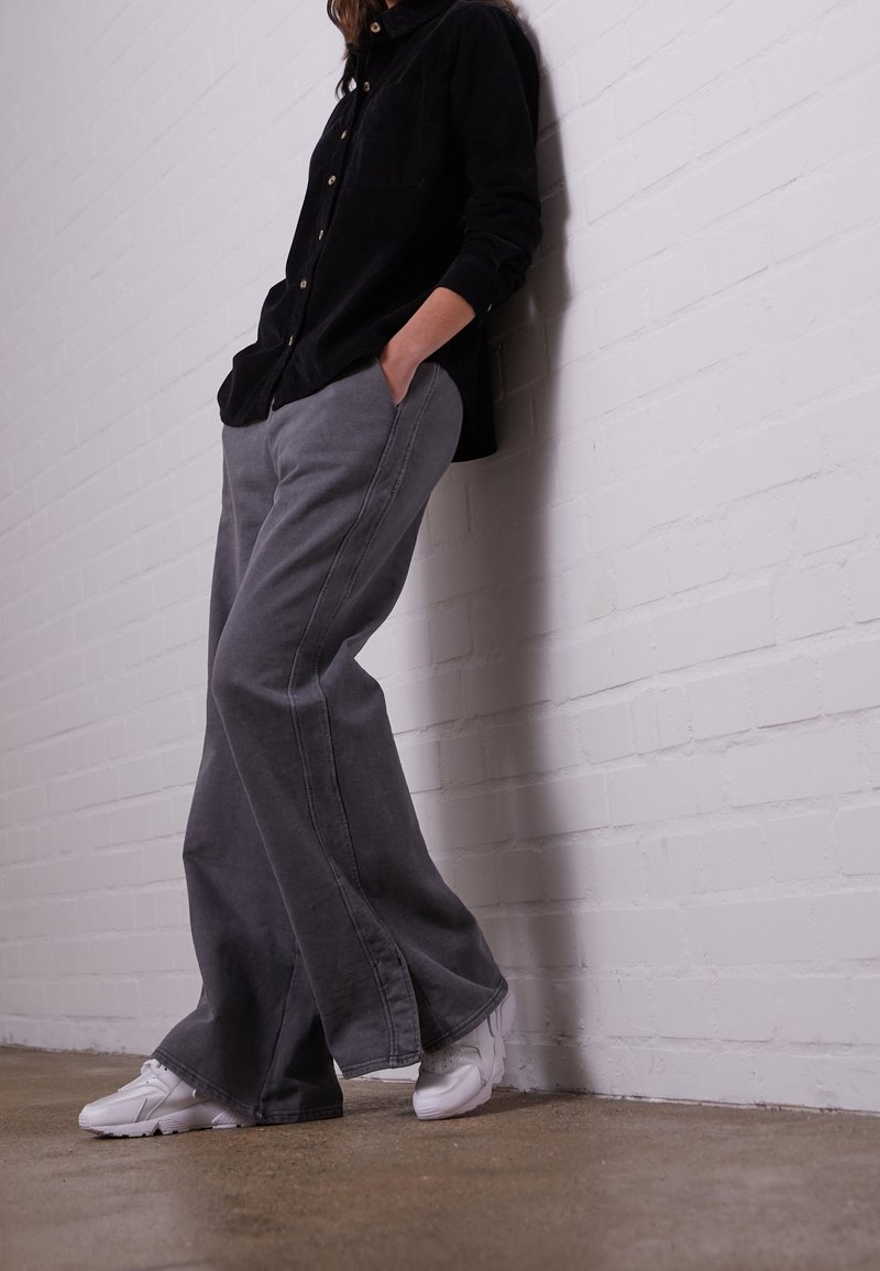 Pantalons en denim gris à jambes larges avec des coutures contrastantes, portés avec une chemise noire à boutons et des baskets blanches, devant un mur en briques blanches.