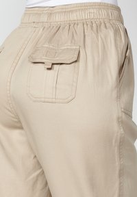 Pantalones cargo beige con cintura elástica, bolsillos laterales y un bolsillo trasero con solapa y detalle de botón. Tejido suave y ligero.