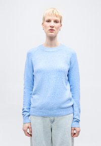 Ξανθός με κοντά μαλλιά, φοράει ανοιχτό μπλε πουλόβερ και τζιν ανοιχτού χρώματος, στέκεται μπροστά σε λευκό φόντο.