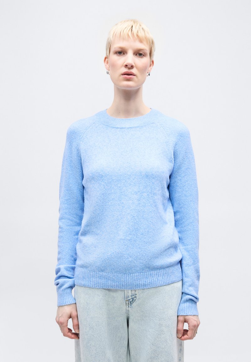 Ξανθός με κοντά μαλλιά, φοράει ανοιχτό μπλε πουλόβερ και τζιν ανοιχτού χρώματος, στέκεται μπροστά σε λευκό φόντο.