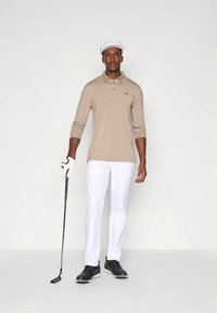 Muž v béžovém polokošili s dlouhým rukávem, bílých kalhotách, bílé čepici, černých golfových botách a rukavici, který drží golfovou hůl, stojící na bílém pozadí.