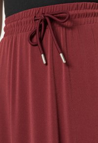 Un pantalon de sport bordeaux avec une taille élastique dotée d'un cordon de serrage et d'accents en métal argenté, présentant un tissu lisse et texturé.