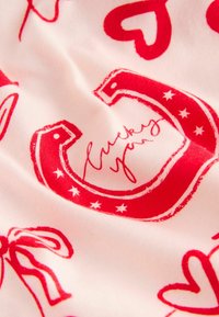 Rødt og hvidt stof med et gentaget mønster af hjerter og hestesko, med teksten "lucky you" og stjerner i en illustrativ stil.