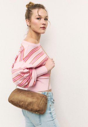 Blonde Frau, die einen rosa gestreiften Pullover und hellblaue Jeans trägt, hält eine braune Wildleder-Handtasche mit einem goldenen Kettenträger über der Schulter.