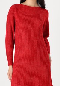 Una modella indossa un maglione di un tessuto strutturato, di un vivace colore rosso, con maniche lunghe e una scollatura ampia, che mette in evidenza il suo motivo a coste e la vestibilità rilassata.
