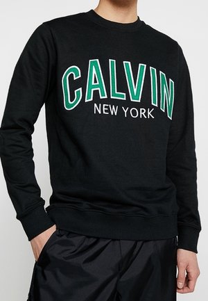 Czarna bluza z długim rękawem z napisem „CALVIN” dużymi zielonymi i białymi literami oraz „NEW YORK” mniejszym białym tekstem na piersi.