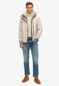 Lys beige vandtætte jakke med høj krave og sort lynlås, båret over en cremefarvet turtleneck, parret med blå jeans og brune støvler.