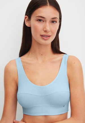 Jonge vrouw met lang donker haar, gekleed in een lichtblauw mouwloos crop top, die licht glimlacht tegen een effen witte achtergrond.