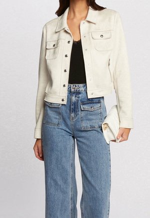 Kvinde iført hvid knapjakke over sort top, højtaljede blå jeans med forlommer, holder en hvid clutch i højre hånd.