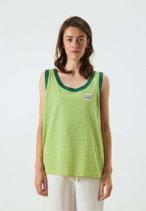 Jonge vrouw die een groen-gele gestreepte mouwloze top met donker groene rand en witte broek draagt, staand tegen een effen achtergrond.