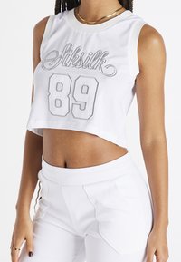 Weiße Cropped-Top mit Netzstruktur, versehen mit dem "Siksilk"-Logo und der Nummer "89" in grauer Stickerei. Kombiniert mit weißen Hosen.