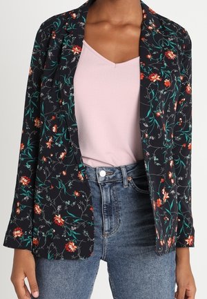 Femme portant un blazer noir à fleurs sur un haut rose clair, assorti à un jean bleu taille haute.