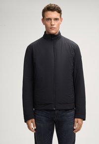 Giacca nera isolante con colletto alto, chiusura frontale con zip e tasche laterali. Il materiale sembra liscio con un design leggero.