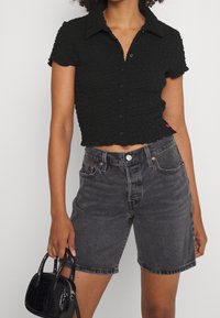 Black textured crop shirt with short sleeves, buttoned front, paired with faded black denim shorts and a small glossy black handbag.