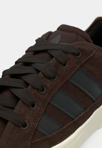 Gros plan d'une basket en daim marron avec des bandes en cuir noir, des lacets assortis marron et une semelle en caoutchouc blanche sur un fond blanc.