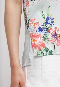 Bloemrijke blouse in lichtblauw, met een verscheidenheid aan gekleurde bloemen (oranje, blauw, groen) en een gladde, lichte stof. Zij-split detail zichtbaar.