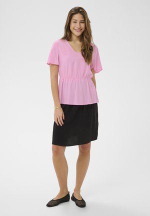 Ženska se smeji, oblečena v roza bluzo s kratkimi rokavi in elastičnim pasom, črno krilo do kolen ter črne tkanih balerinke, stoji pred enobarvnim ozadjem.