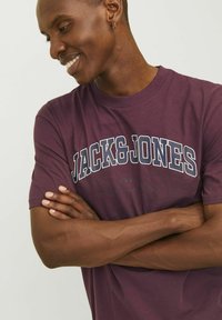 Camiseta de algodón marrón con mangas cortas, que presenta texto impreso en blanco y azul "JACK&JONES" y "EST. 1990 WORLDWIDE" en el pecho.