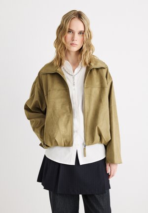 Mladá žena s vlnitými blond vlasy, oblečená v olivově zelené bundě na zip, bílém košili na knoflíky, stříbrném řetízku, černé sukni a tmavých kalhotách.