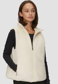 Biała kamizelka z polaru z wysokim kołnierzem, zamkiem błyskawicznym z przodu i kieszeniami po bokach, noszona na czarnej bluzie z długim rękawem. Miękka faktura i luźny krój.