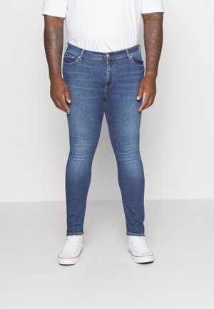 Homme portant un jean bleu ajusté, des baskets blanches et un t-shirt blanc, debout devant un fond clair uni.
