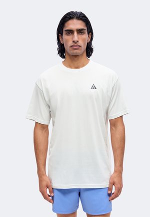 Mand med mellemlangt sort hår iført en hvid ACG-logo t-shirt og lyseblå shorts, der står mod en ensfarvet baggrund.