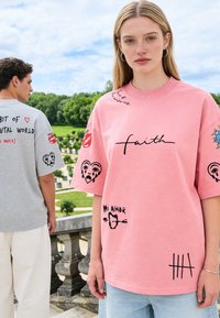 Jeune femme en chemise rose oversize avec "faith" et dessins, debout à côté d'un homme en chemise grise avec texte et graphiques, en extérieur près d'une balustrade en pierre.