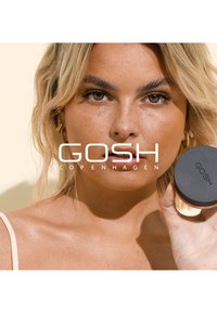 Czarny pojemnik kosmetyczny trzymany w ręce, z napisem "GOSH" na wieczku. Przedstawia kobietę z żółtozłotymi kolczykami w kształcie kół i blond włosami.