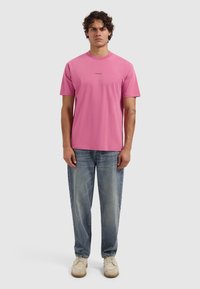 Junger Mann steht mit einem rosa T-Shirt mit dem Aufdruck "PURE PATH", blauen Jeans, beigen Schuhen und einem Tattoo am linken Unterarm.