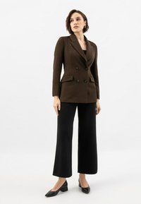 Blazer marron à double boutonnage avec des revers en pointe, deux poches avant et un pantalon noir à jambes larges, associé à des chaussures à talons noires.