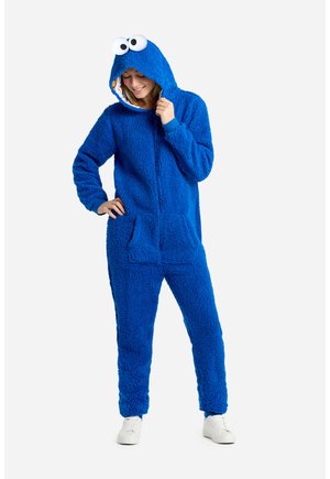 Combinaison en polaire bleue avec zip sur le devant, grande capuche avec des yeux globuleux, et poches latérales. Tissu doux et texturé, coupe décontractée.