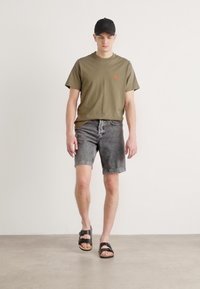 T-shirt vert à manches courtes, short en denim gris délavé et sandales noires. Tenue décontractée au design minimaliste et aux couleurs neutres.