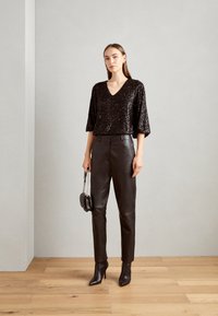 Schwarzes Pailletten-Top mit V-Ausschnitt und lockeren Ärmeln, kombiniert mit engen schwarzen Lederhosen und spitzen Ankle-Boots. Kleine schwarze Schultertasche.