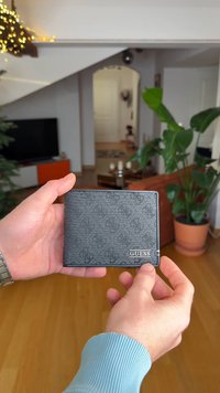 Portafoglio Guess nero con finitura strutturata, caratterizzato da un logo discreto e un bordo liscio. Forma rettangolare compatta, tenuto in mano.