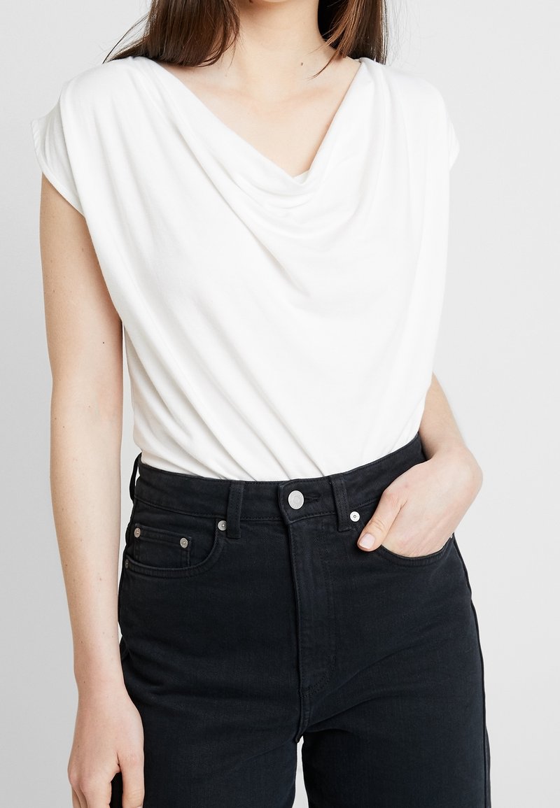 Top blanco con cuello de cowl en tejido ligero, de manga corta. Combinado con jeans negros de tiro alto que cuentan con bolsillos frontales y herrajes plateados.